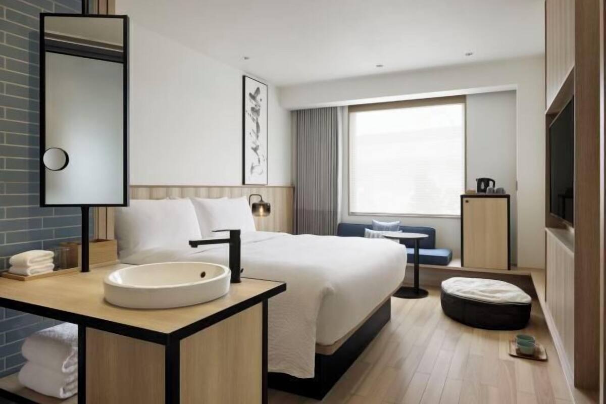A modern hotel room with a large bed, a round bathroom sink, a bedside table, a large window, and a TV.