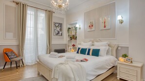 A bedroom with a large bed, a nightstand, a chair, a chandelier, and wall art.