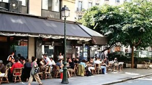 An outdoor cafe with tables and chairs, a street lamp, and buildings in the background.