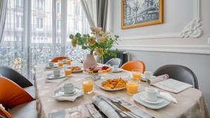 A breakfast setting with a table set for two, featuring croissants, pastries, and orange juice. A vase with flowers is on the table, and a framed picture hangs on the wall.