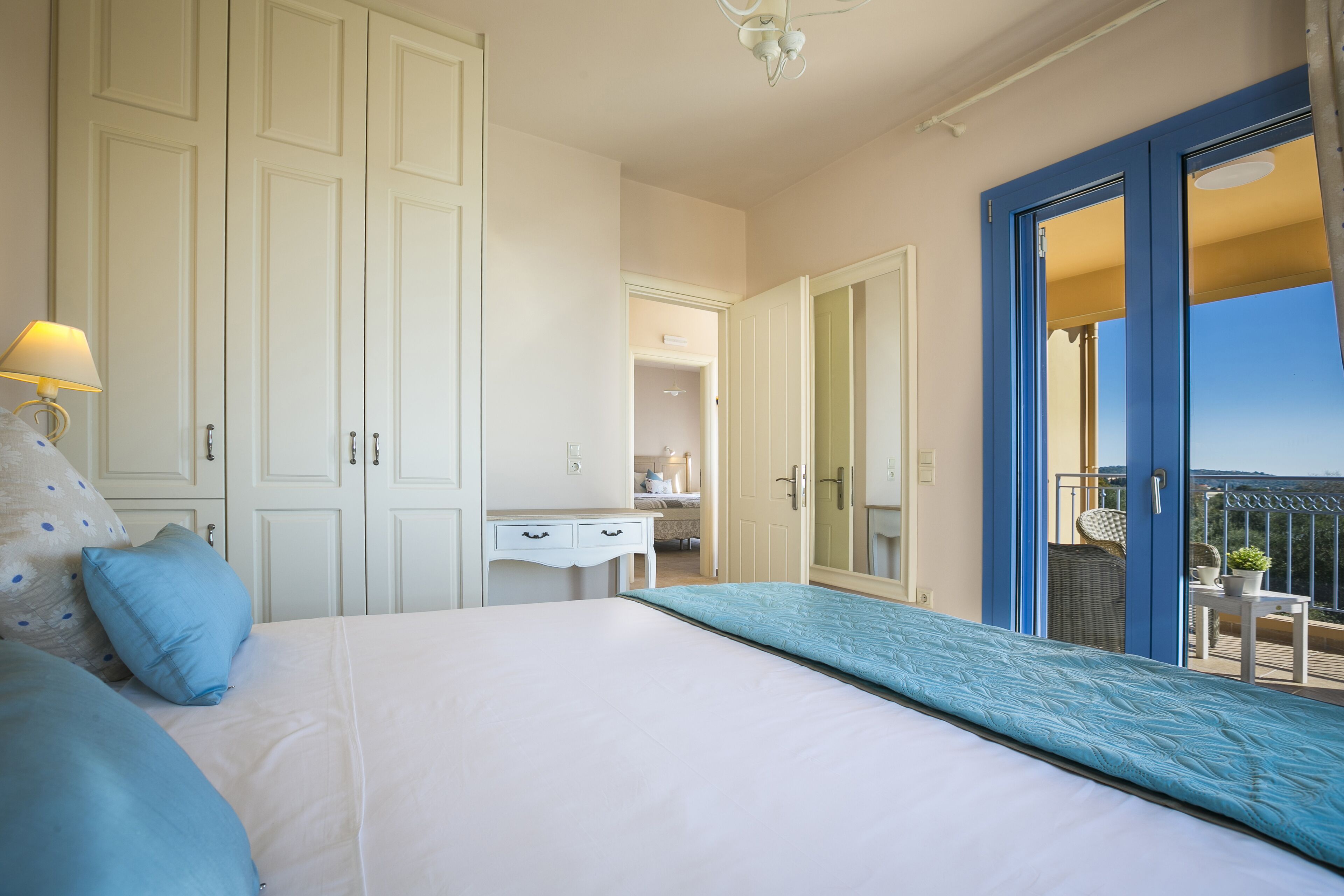 A bedroom with a large bed, a desk, and a wardrobe. There is a balcony with a view.