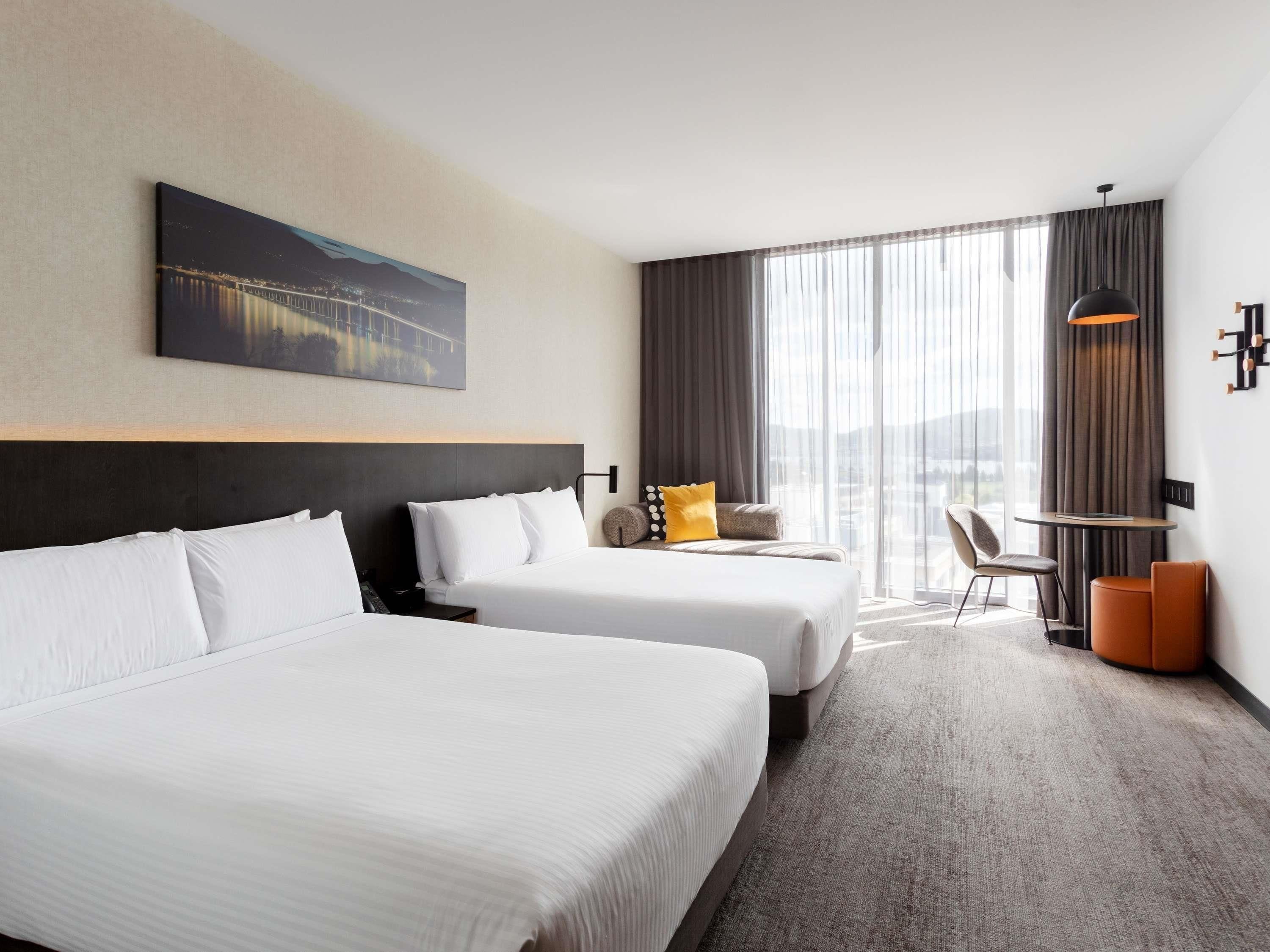 A hotel room with two beds, a desk, a chair, a large window with curtains, and a painting on the wall.