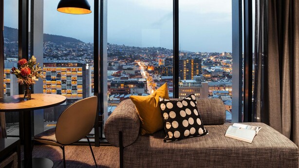 A modern room with a large window, a round table, a sofa with patterned cushions, and a hanging pendant light.