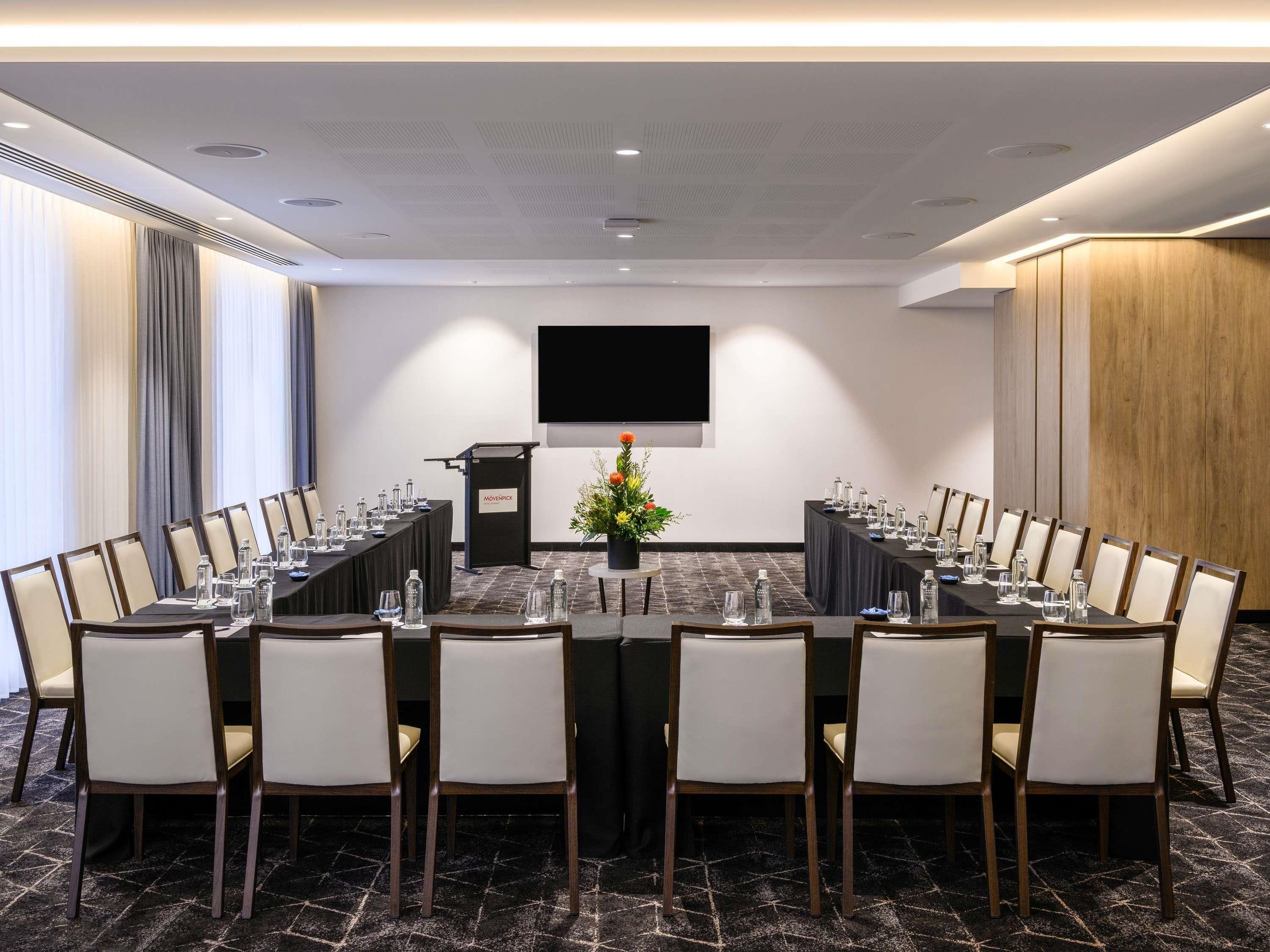 A conference room with a long table, chairs, a podium, a TV screen, and a flower arrangement.
