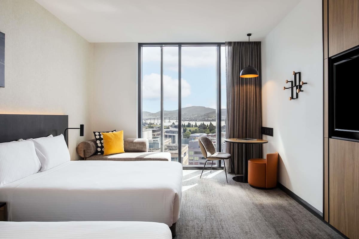 Een hotelkamer met twee bedden, een groot raam met uitzicht op de stad, een tafeltje met een lamp en een aan de muur bevestigde televisie.