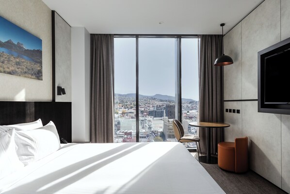 Una habitación de hotel moderna con una cama grande, una mesa redonda, una silla, un televisor montado en la pared y vistas a la ciudad a través de amplias ventanas.