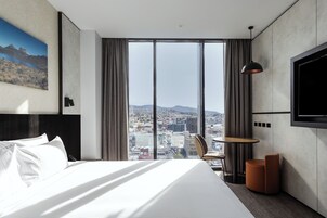A modern hotel room with a large bed, a round table, a chair, a TV mounted on the wall, and a city view through large windows.