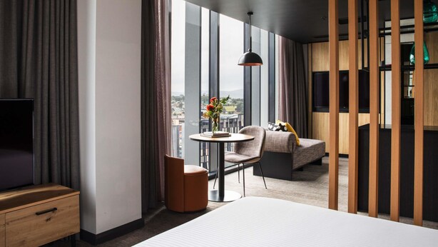 Et moderne hotelværelse med en stor seng, et lille rundt bord med en stol, et fjernsyn og udsigt over byen gennem store vinduer.