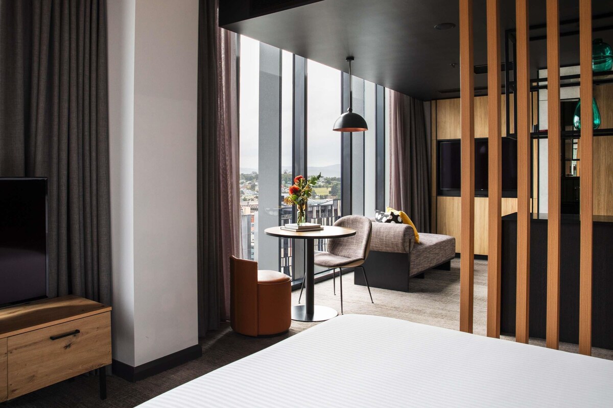 Una habitación de hotel moderna con una cama grande, una mesita redonda con una silla, televisión y vistas a la ciudad a través de amplios ventanales.