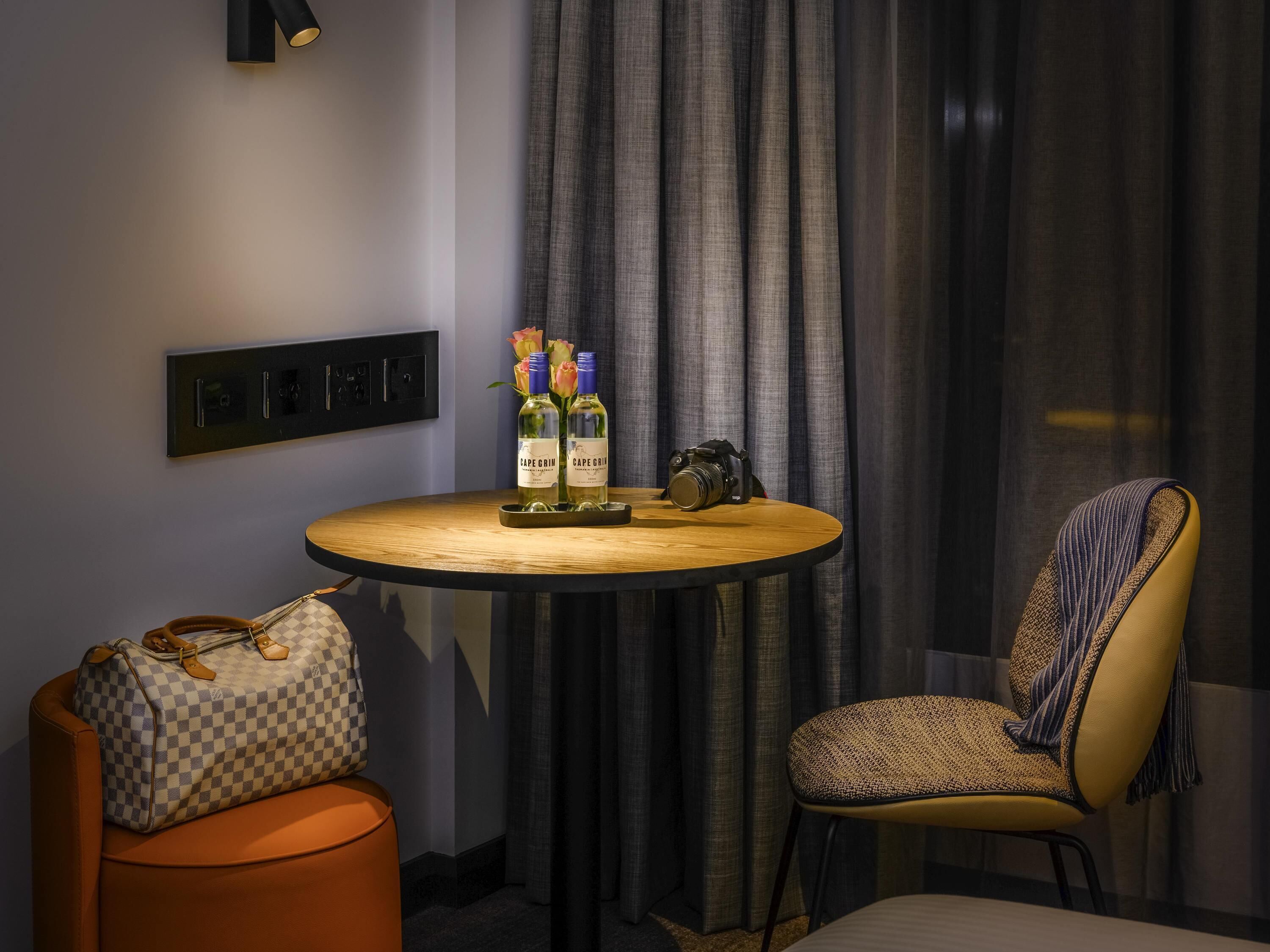 A hotel room with a round wooden table, two bottles of Cape Grain wine, a camera, a checkered handbag, and a patterned armchair.