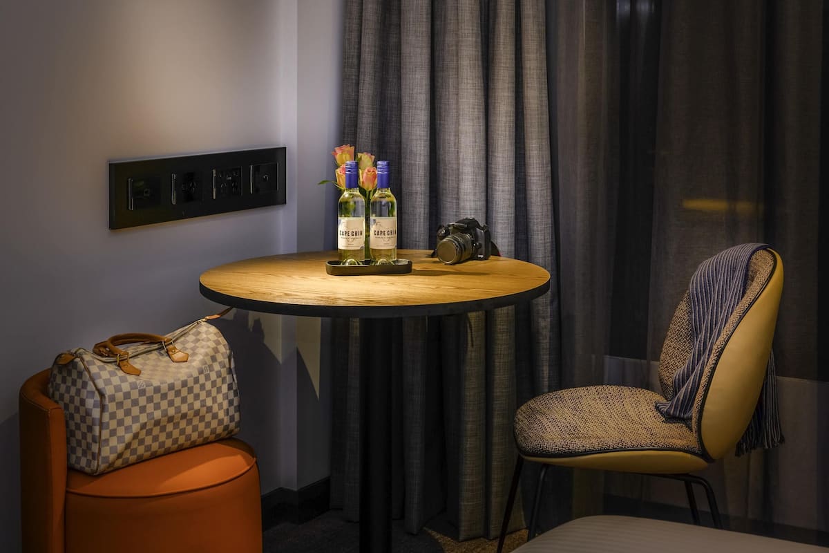 A hotel room with a round wooden table, two bottles of Cape Grain wine, a camera, a checkered handbag, and a patterned armchair.