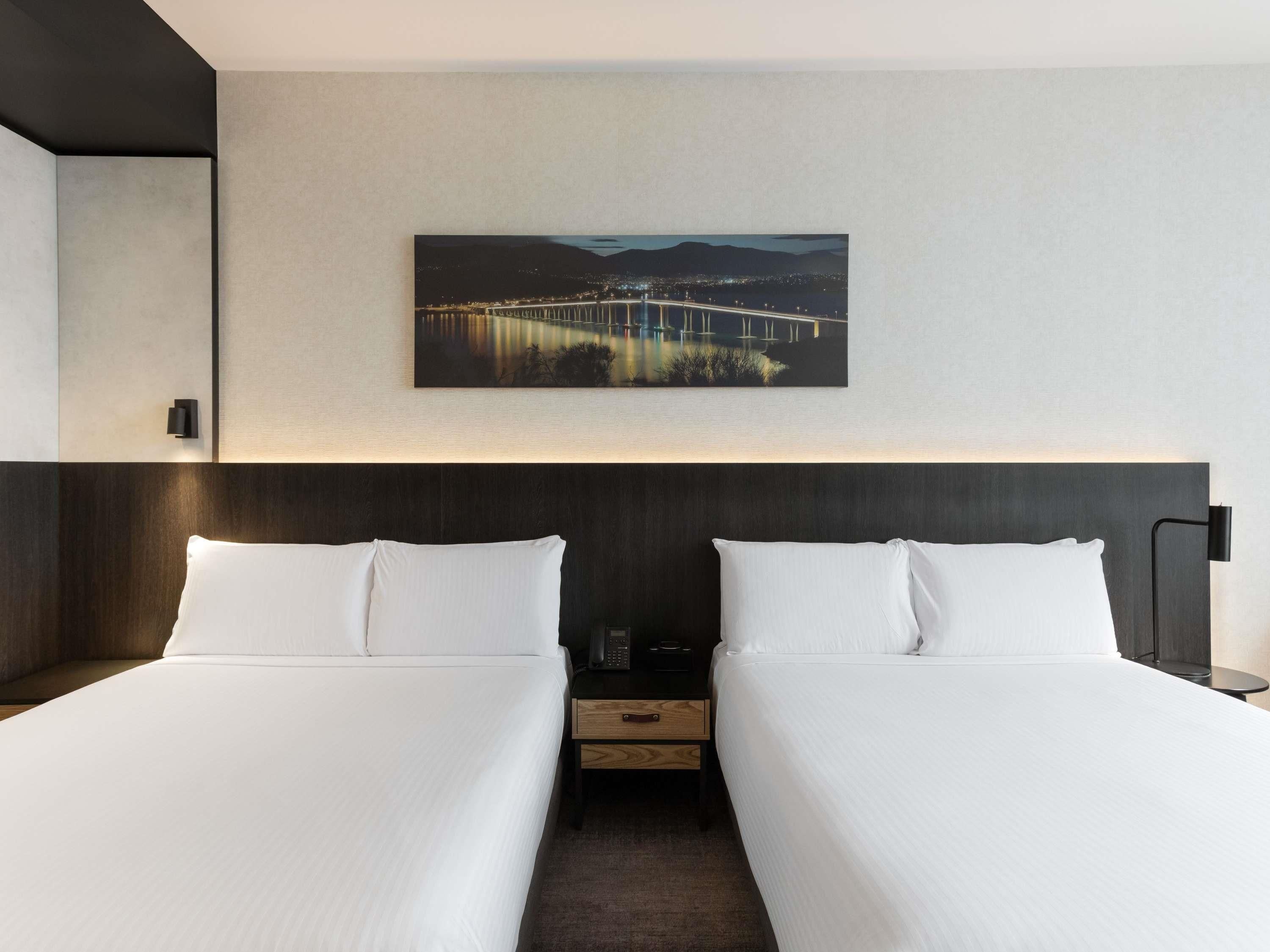 A hotel room with two beds, a nightstand, a lamp, and a painting of a bridge.