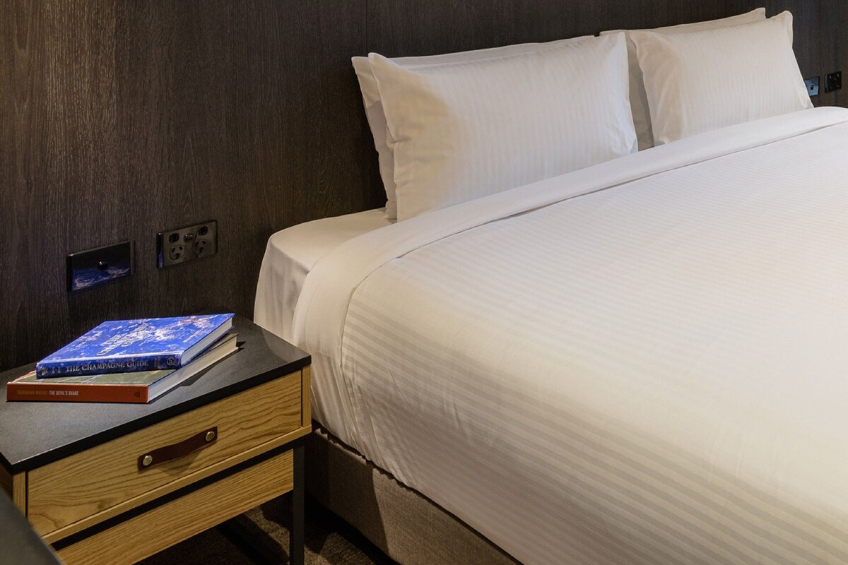 Una cama bien hecha con sábanas blancas, una mesita de noche de madera con un libro y un enchufe, y una pared de madera oscura.