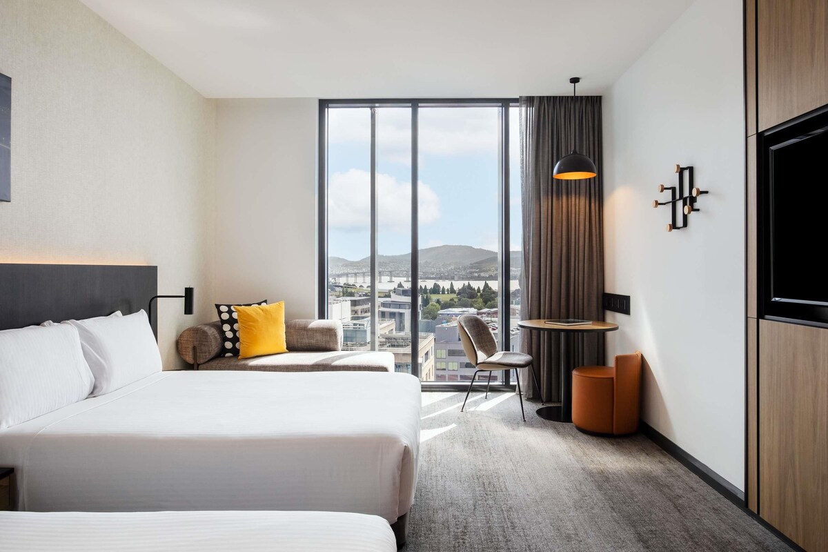 Een hotelkamer met twee bedden, een groot raam met uitzicht op de stad, een tafeltje met een lamp en een aan de muur bevestigde televisie.
