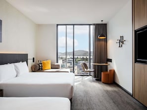 Habitación de hotel con dos camas, un ventanal con vista a la ciudad, una mesita con lámpara y un televisor montado en la pared.