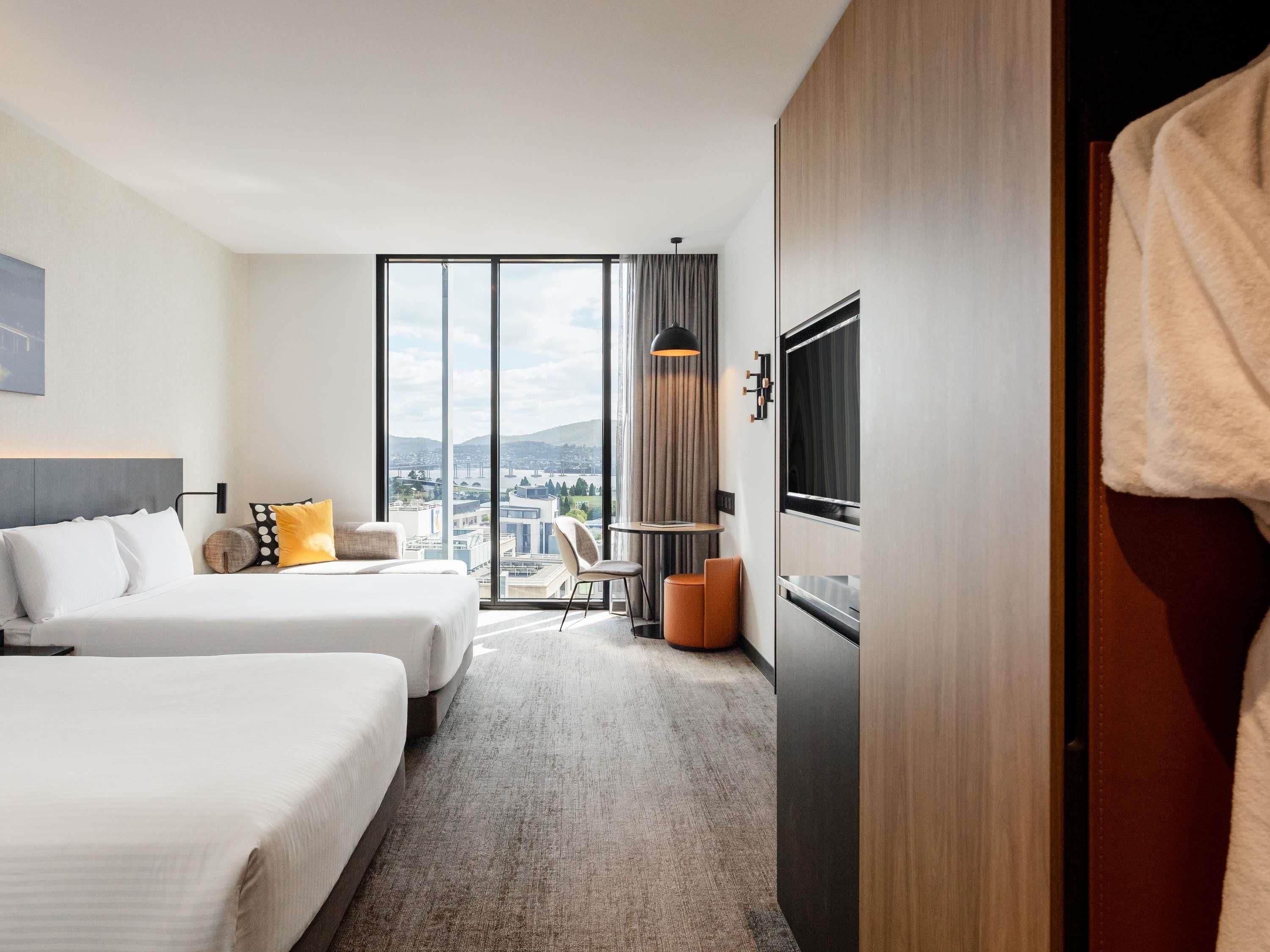 A hotel room with two beds, a desk, a chair, a TV, and a large window with a city view.