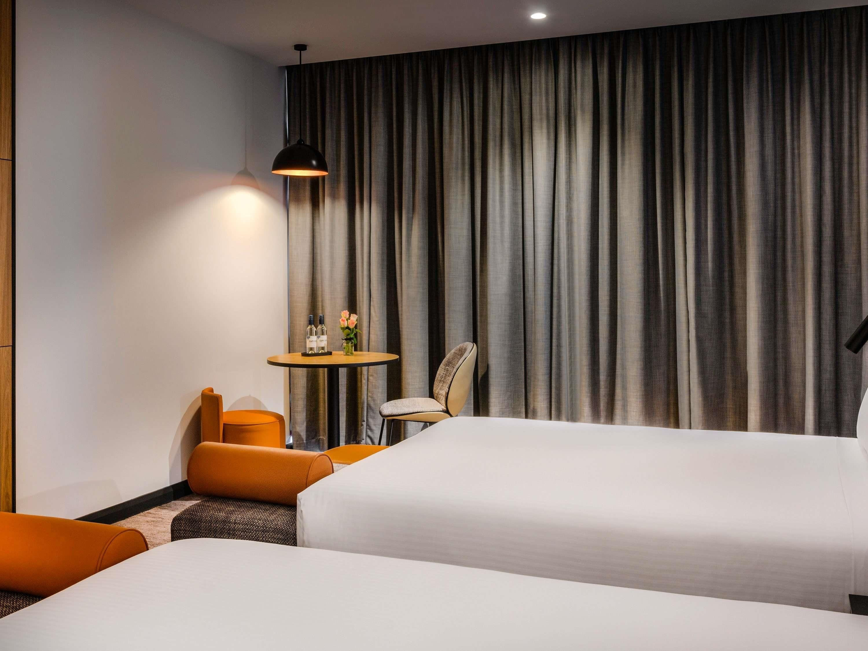 A hotel room with two beds, a small table with a lamp, and a chair.