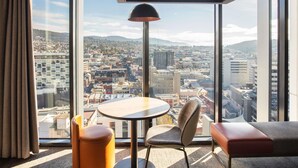 A modern hotel room with a large window, a round table, a cushioned chair, and a sofa.