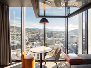 A modern hotel room with a large window, a round table, a cushioned chair, and a sofa.