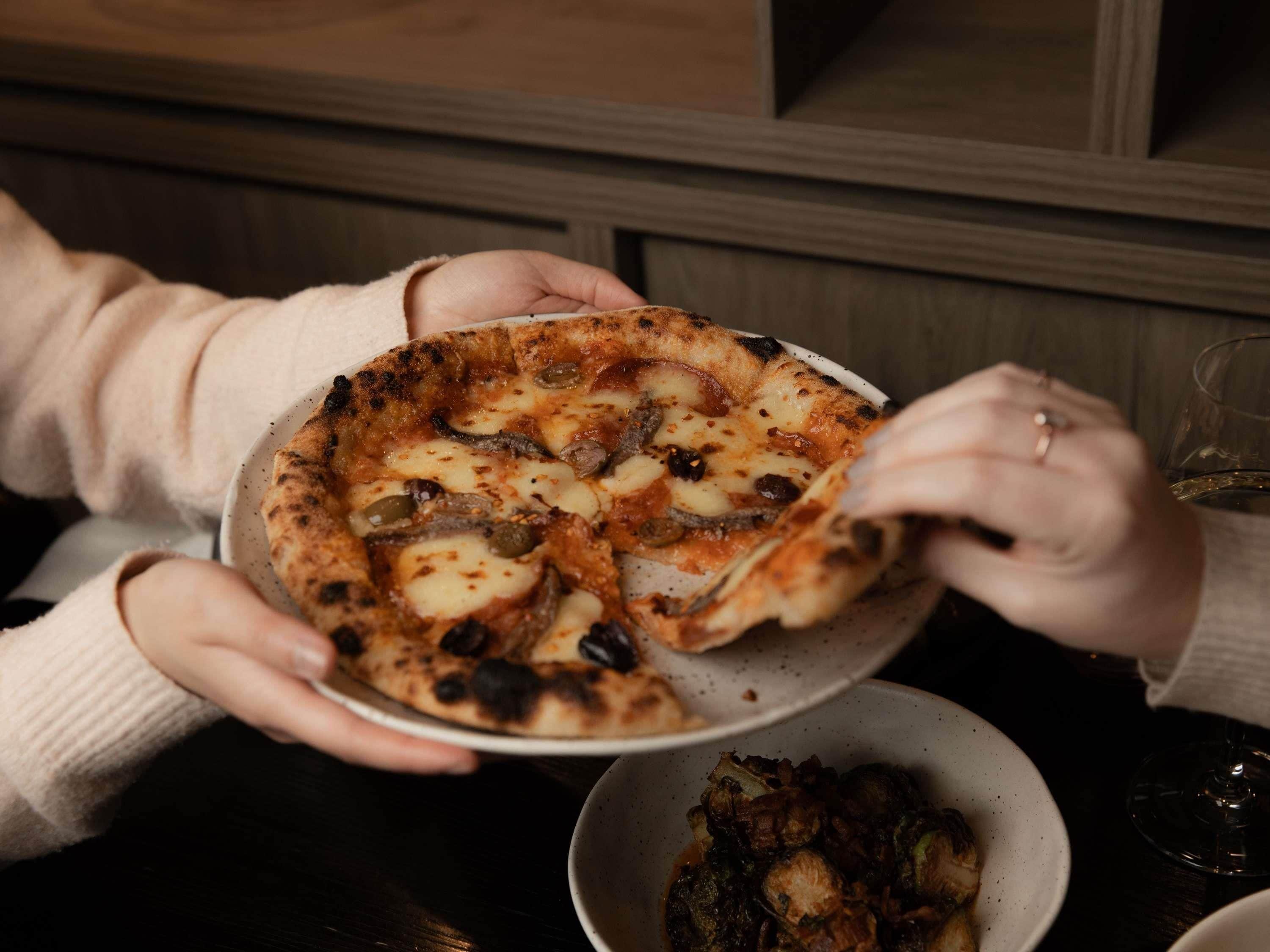 Eine Person hält eine Pizza, von der ein Stück herausgehoben wird, zusammen mit einem Brüsseler Rosenkohl-Boul und einem Weinglas auf einem Tisch.