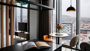 A modern hotel room with a large window, a desk with a book, a chair, a small table with a vase, and a television.