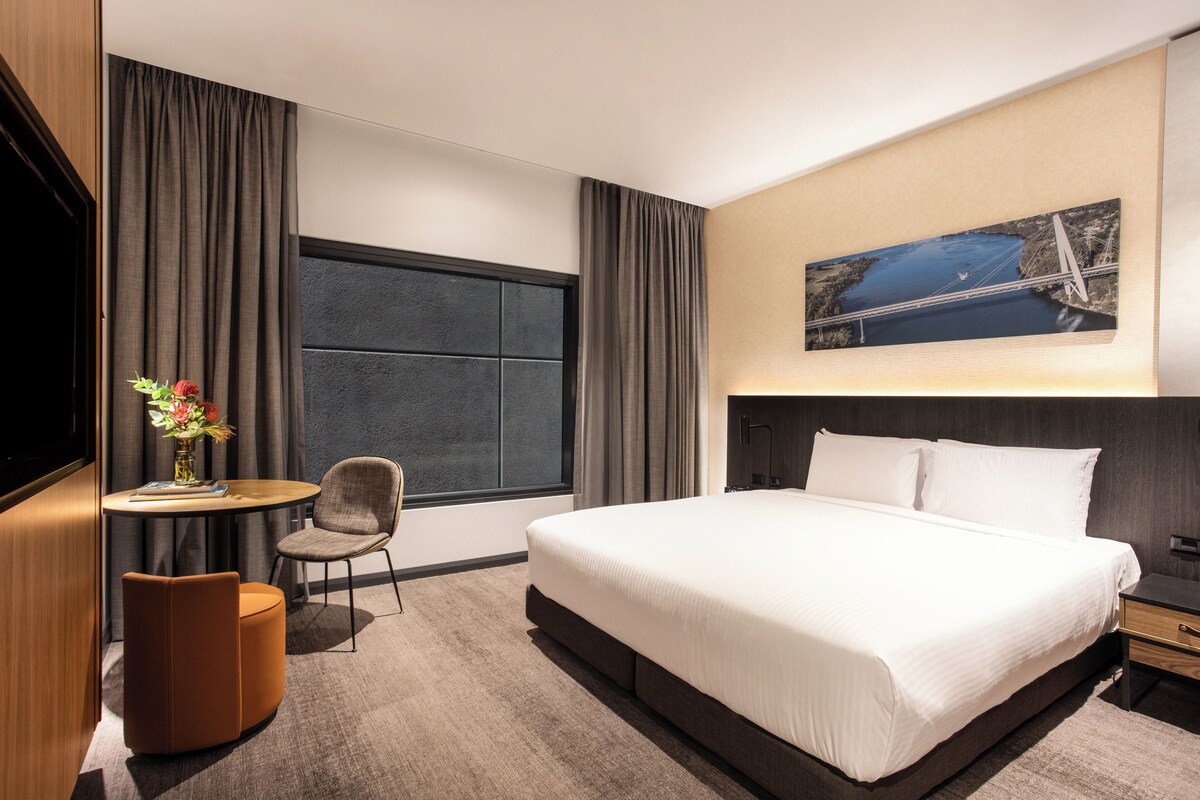 Una habitación de hotel moderna con una cama grande, una mesita redonda pequeña con una silla, un televisor y un cuadro de un puente en la pared.