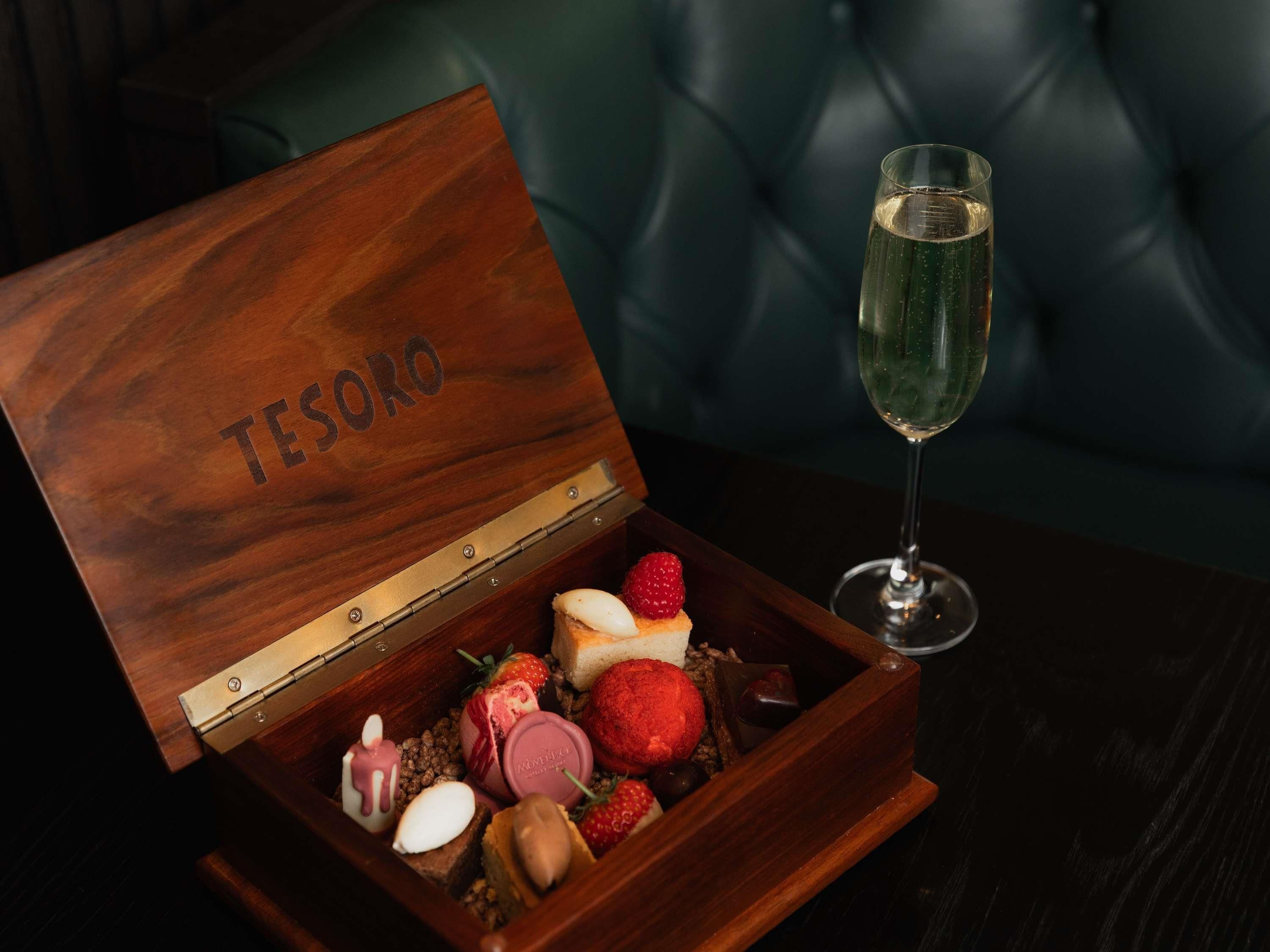 A wooden box with the word 'TESO' on it, containing various chocolates and a glass of champagne.