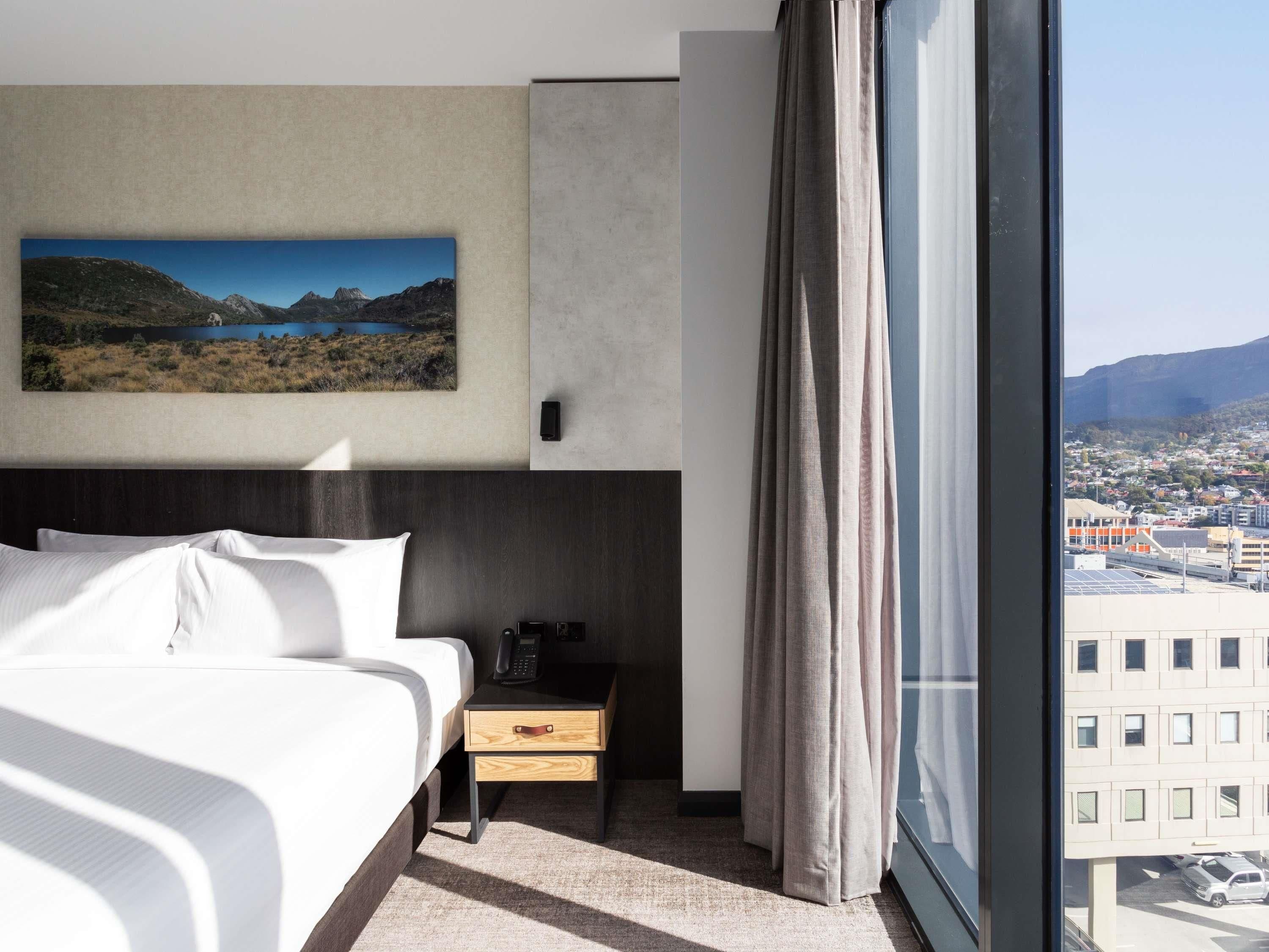 A hotel room with a bed, a bedside table with a phone, a painting of a landscape, and a view of a city through large windows.