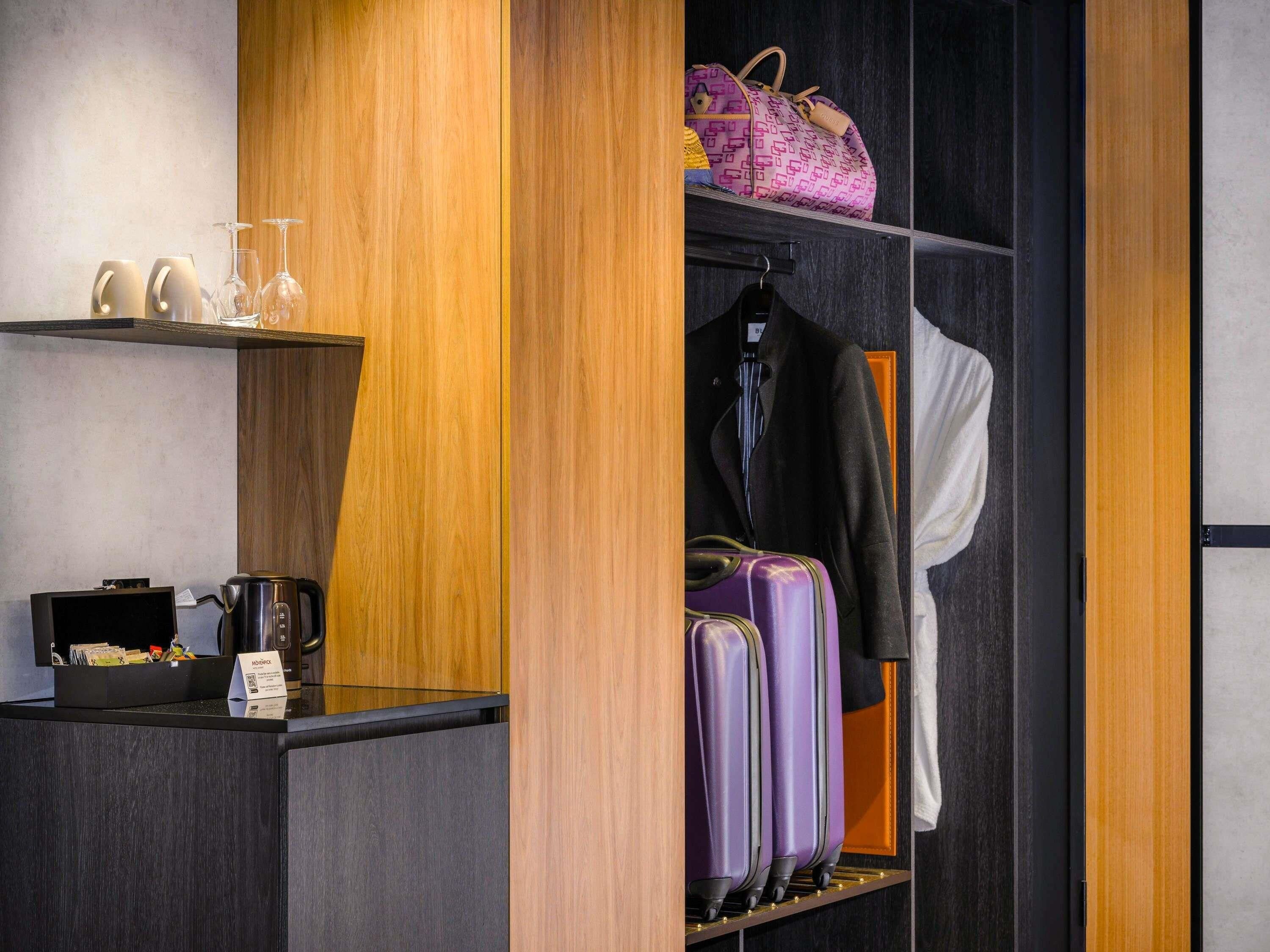 A hotel room with a wooden reception desk, a coffee machine, a shelf with wine glasses, a hanging coat, and a rack with suitcases.