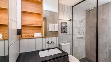 A modern bathroom with a black countertop, a white sink, a toilet, a shower with a glass door, and a wooden vanity with shelves.