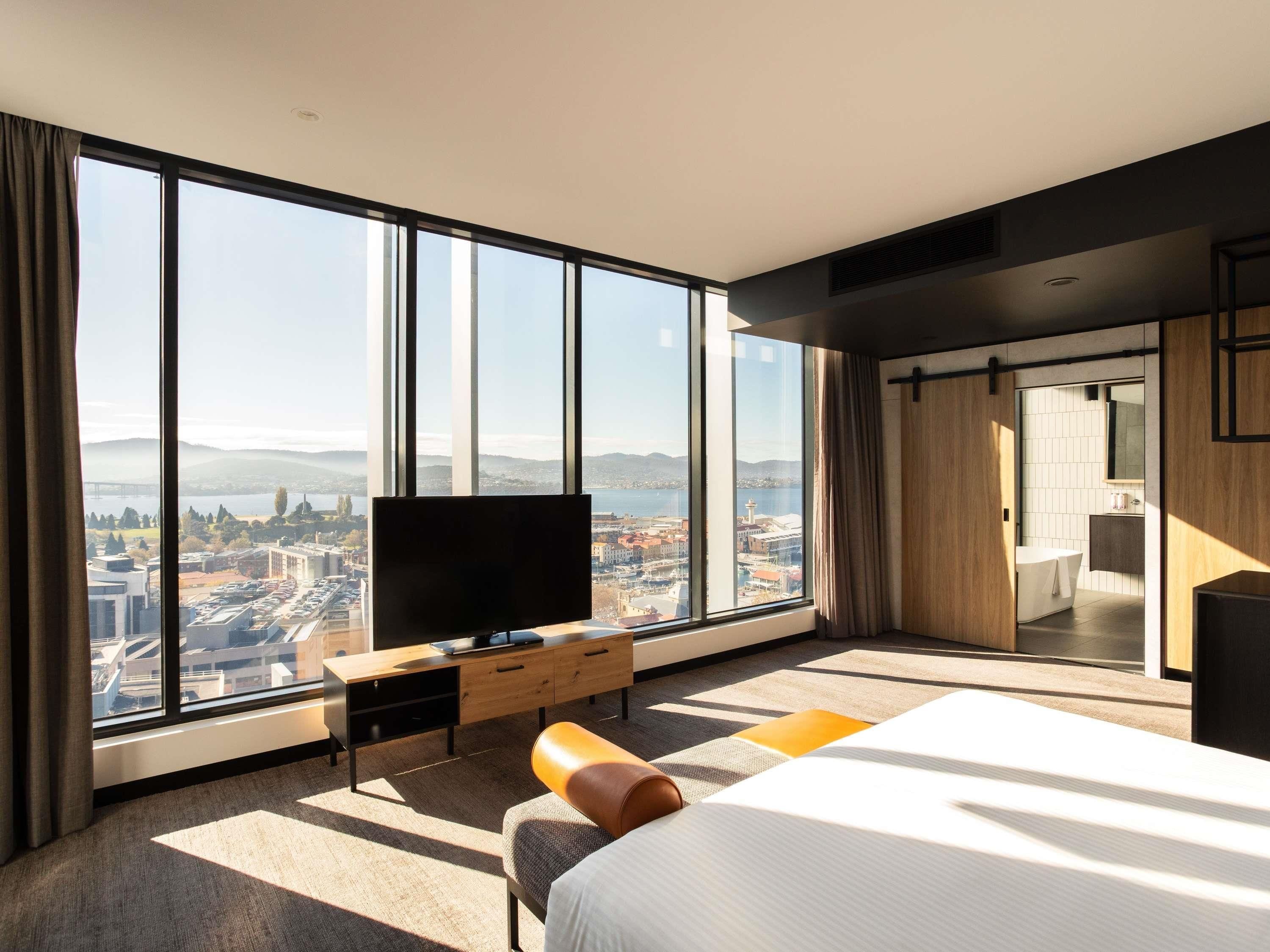 Una habitación de hotel moderna con un ventanal grande, un televisor de pantalla plana, una cama, un escritorio y un baño visible a través de una puerta abierta.