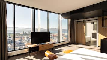 Una habitación de hotel moderna con un ventanal grande, un televisor de pantalla plana, una cama, un escritorio y un baño visible a través de una puerta abierta.