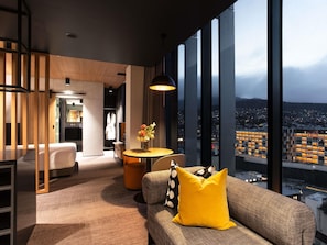 Una habitación de hotel moderna con un ventanal grande, un sofá con un cojín amarillo, una mesa redonda con un florero y una partición de madera.