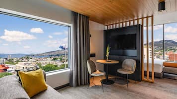 A modern hotel room with a large window, a sofa, a small table with chairs, and a flat-screen TV mounted on the wall.