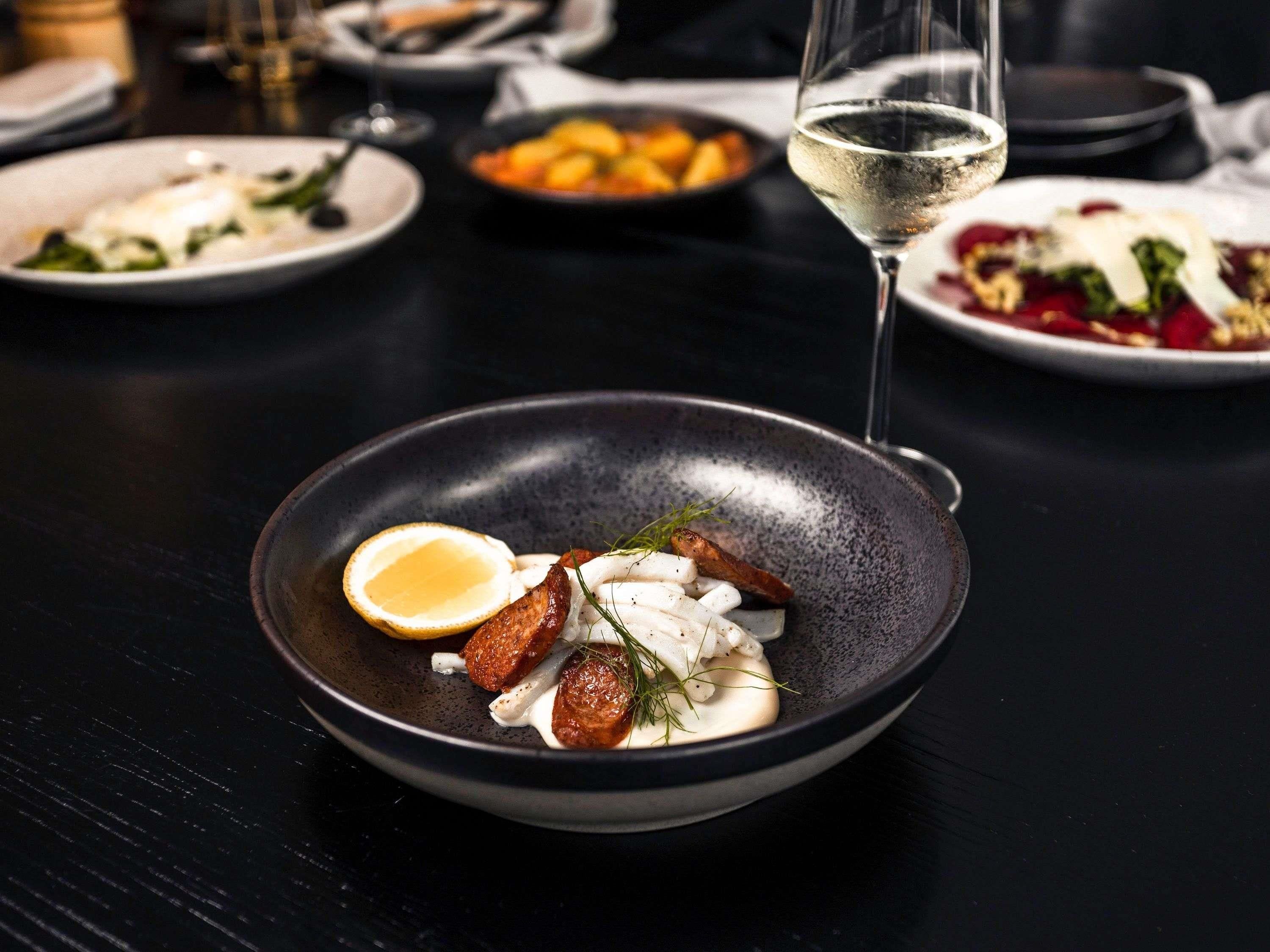 Une table à manger avec un bol noir contenant de la nourriture, un verre de vin blanc et, au fond, des assiettes avec différents plats.