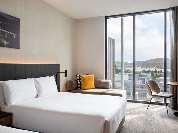 Habitación de hotel con una cama grande, zona de estar con una silla, una mesa y vistas a un paisaje urbano a través de amplios ventanales.