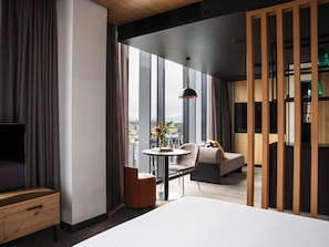 Una habitación de hotel moderna con una cama, una mesita con un jarrón, una silla, un televisor y un ventanal con vistas a la ciudad.