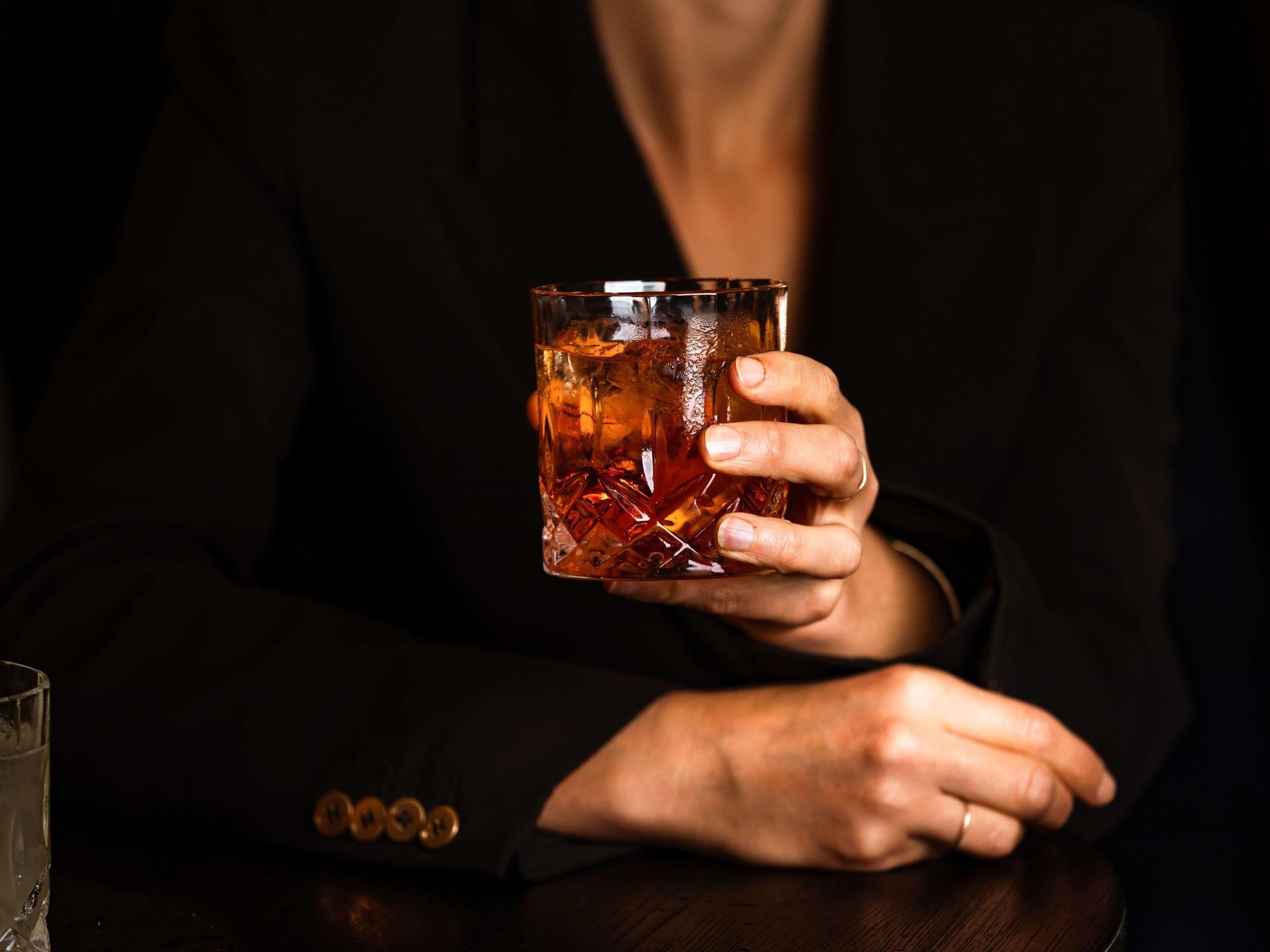 A person holding a glass with a dark liquid, wearing a dark suit, seated at a wooden table.