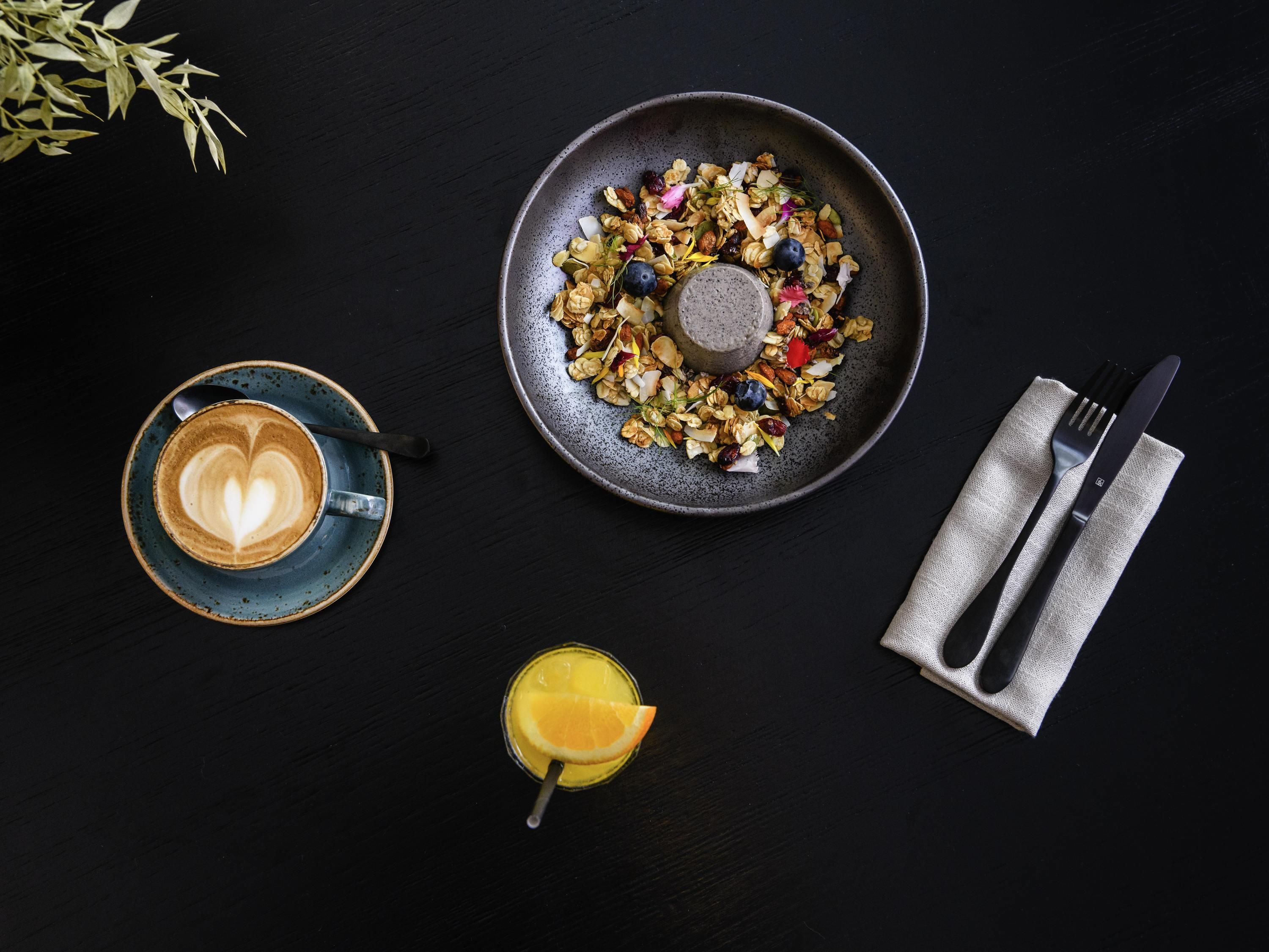 A breakfast setting with a plate of granola, a cup of coffee with latte art, a glass of orange juice, and cutlery on a dark table.