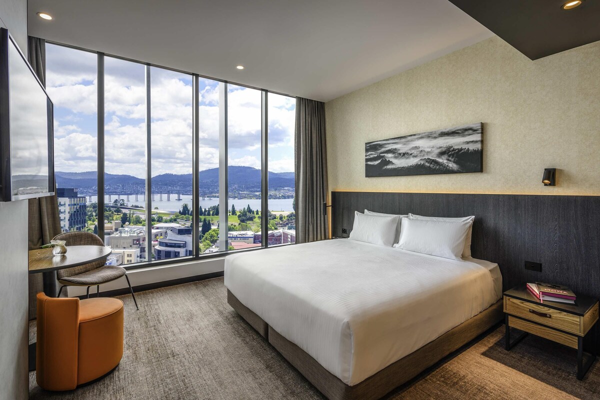 Hotelkamer met een groot bed, een bureau, een stoel en een stadszicht door grote ramen.