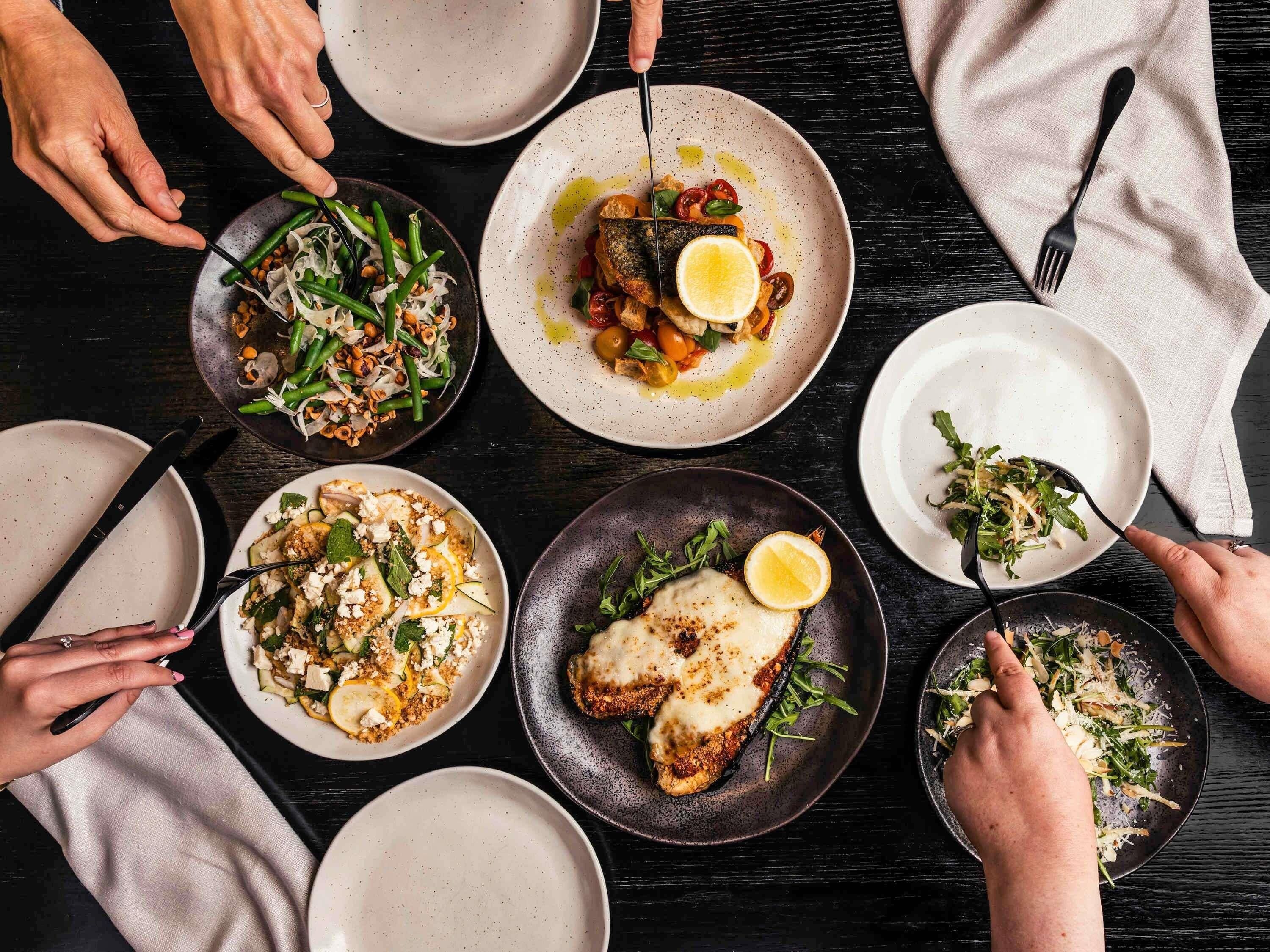 A dining table with multiple plates of food, utensils, and hands reaching for the plates.