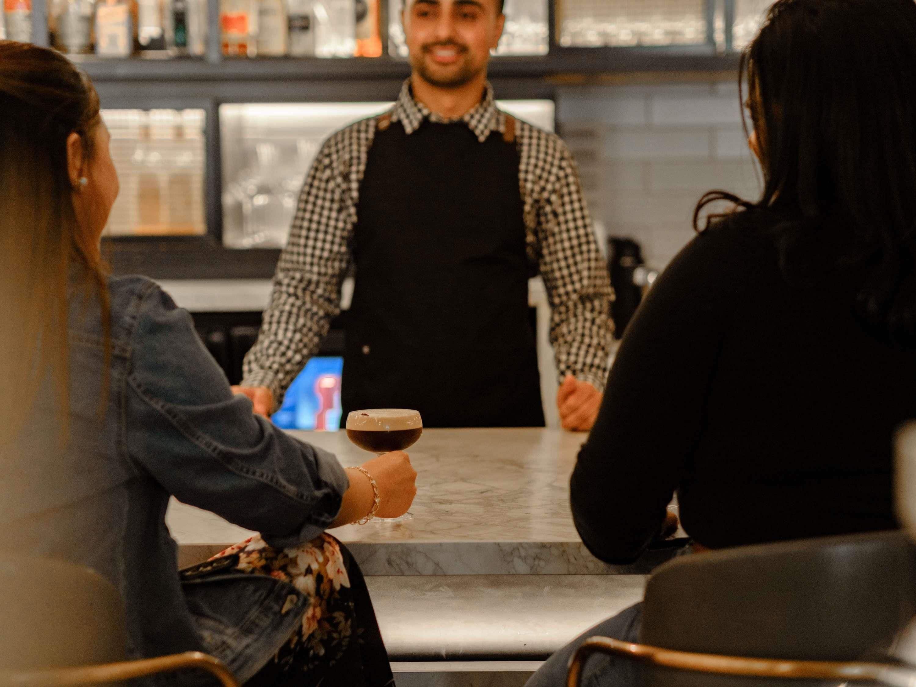 A bar setting with a counter, a person standing behind it, and two seated individuals.