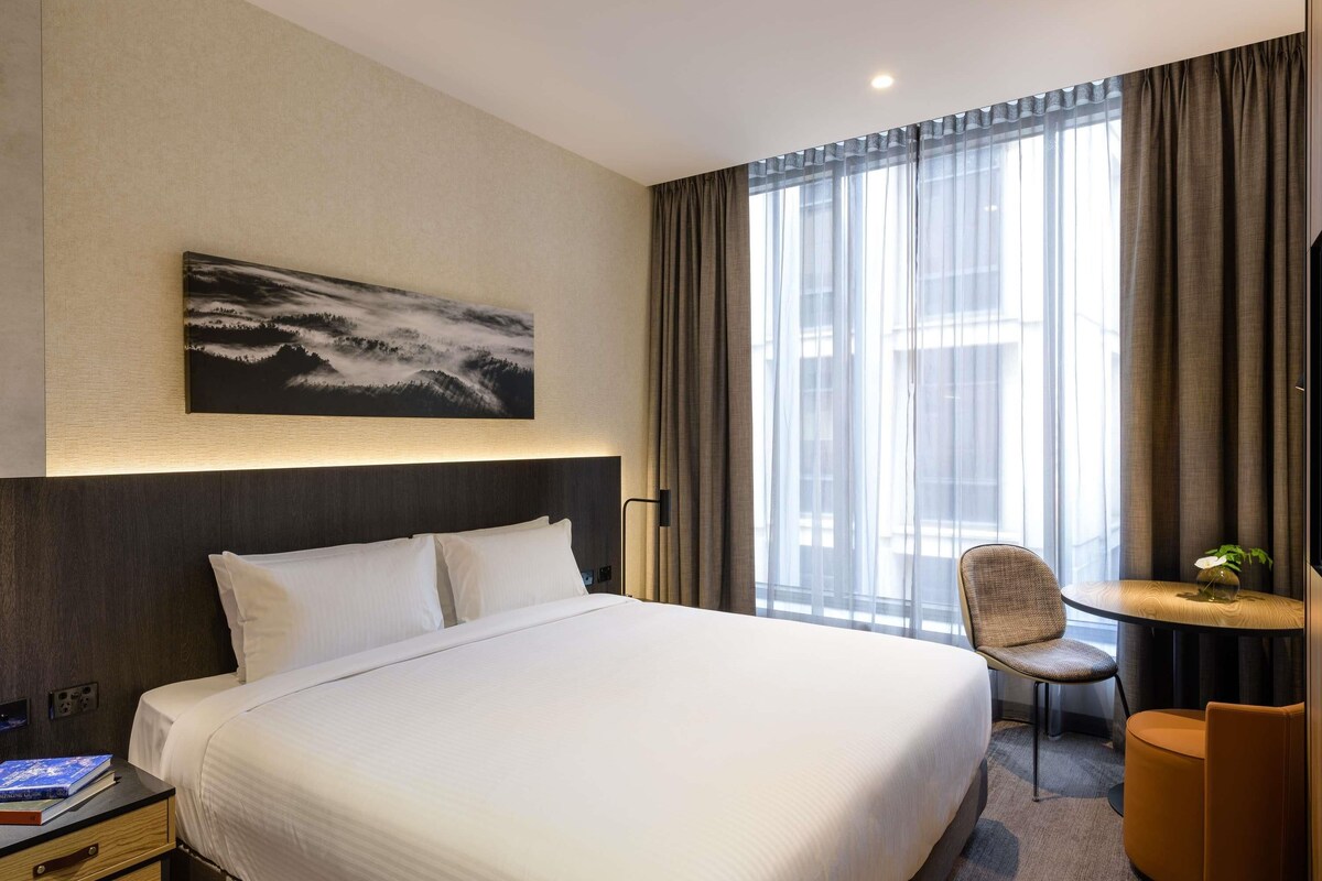 Een hotelkamer met een groot bed, een stoel, een klein tafeltje en een schilderij aan de muur.