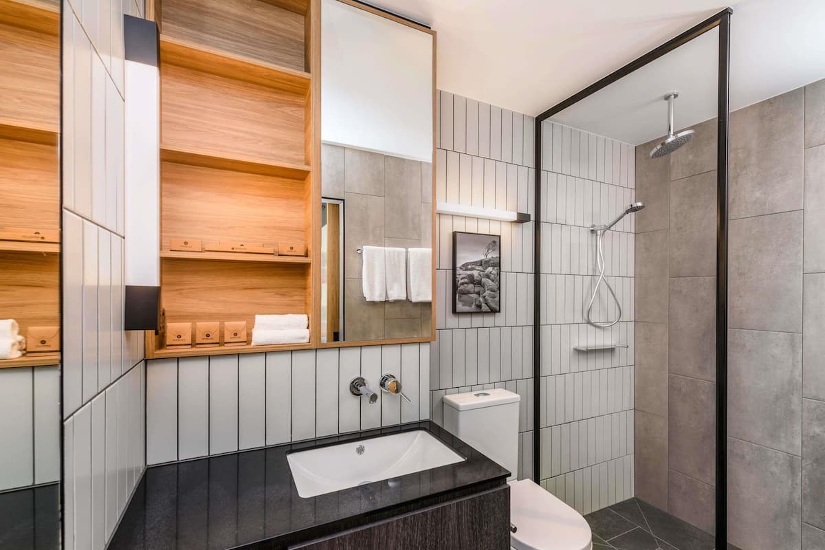 Een moderne badkamer met een zwart aanrecht, een witte wastafel, een toilet, een douche met glazen deur en een houten wastafelmeubel met planken.