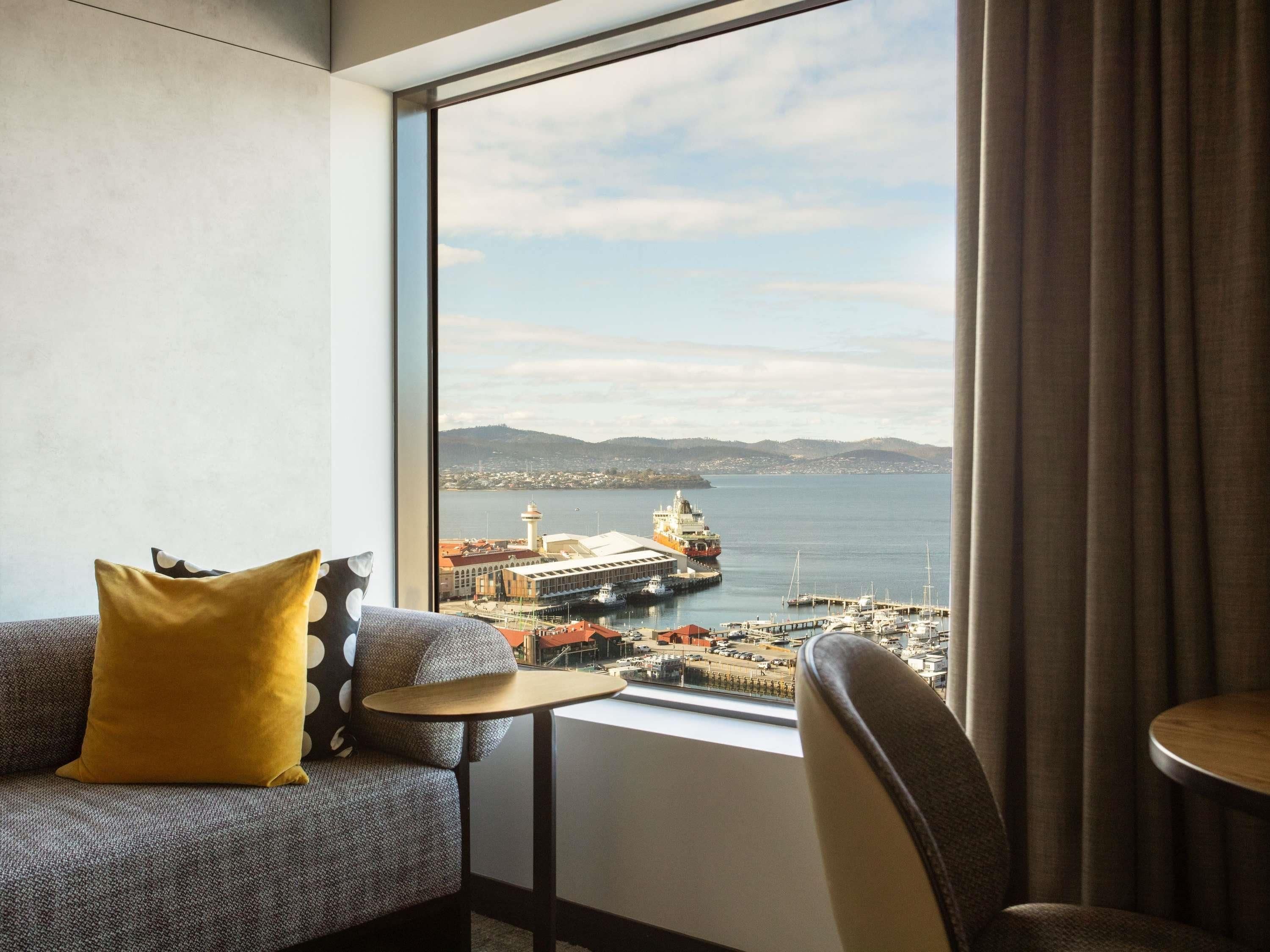 A hotel room with a large window offering a view of a harbor, a lighthouse, and distant hills.