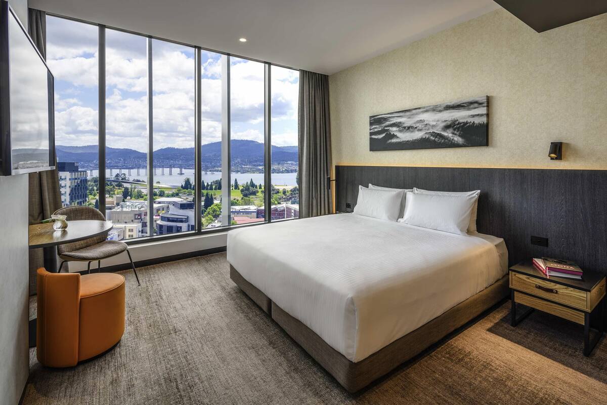 Hotelkamer met een groot bed, een bureau, een stoel en een stadszicht door grote ramen.