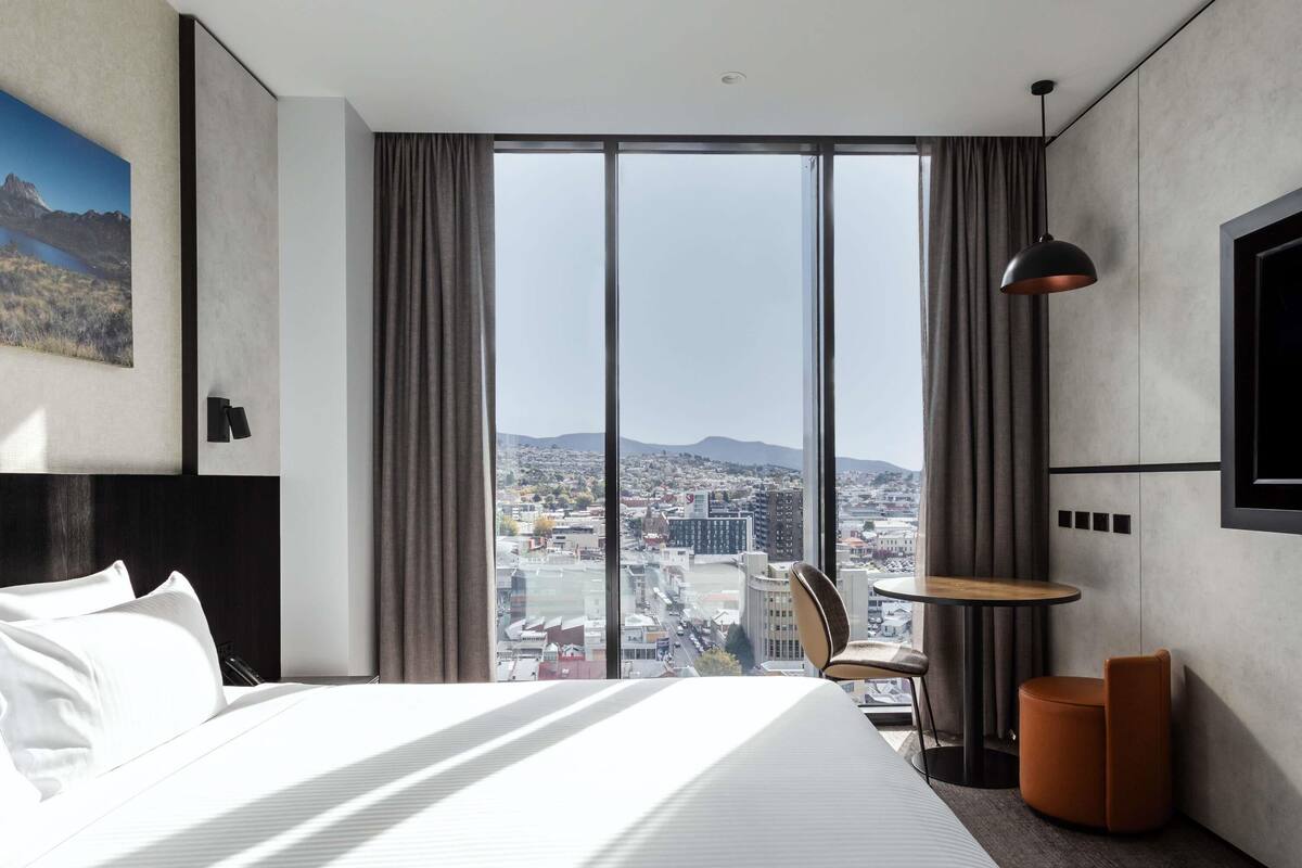 Een hotelkamer met een groot bed, een kleintje tafeltje, een stoel, een televisie en uitzicht over de stad.