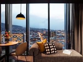 A modern room with a large window, a round table, a sofa with patterned cushions, and a hanging pendant light.