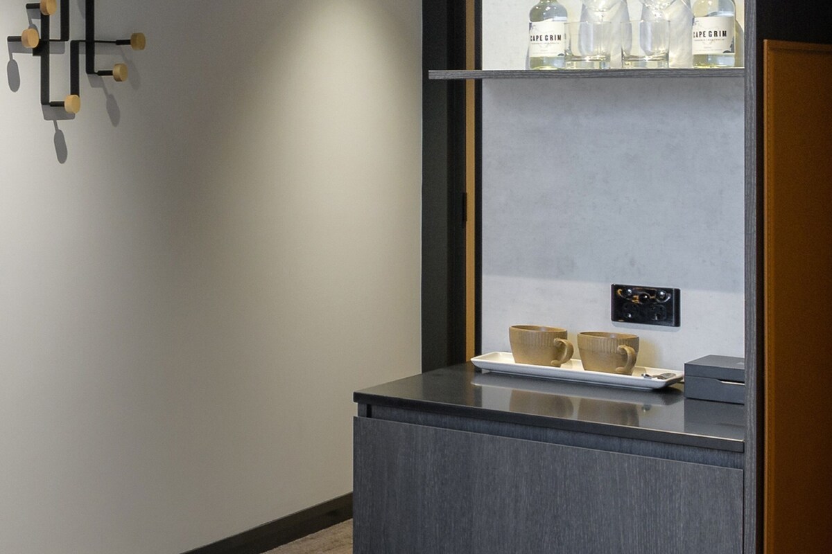 Una habitación de hotel moderna con una consola gris oscuro, dos tazas color beige y una botella de vino Cape Grim en una estantería.