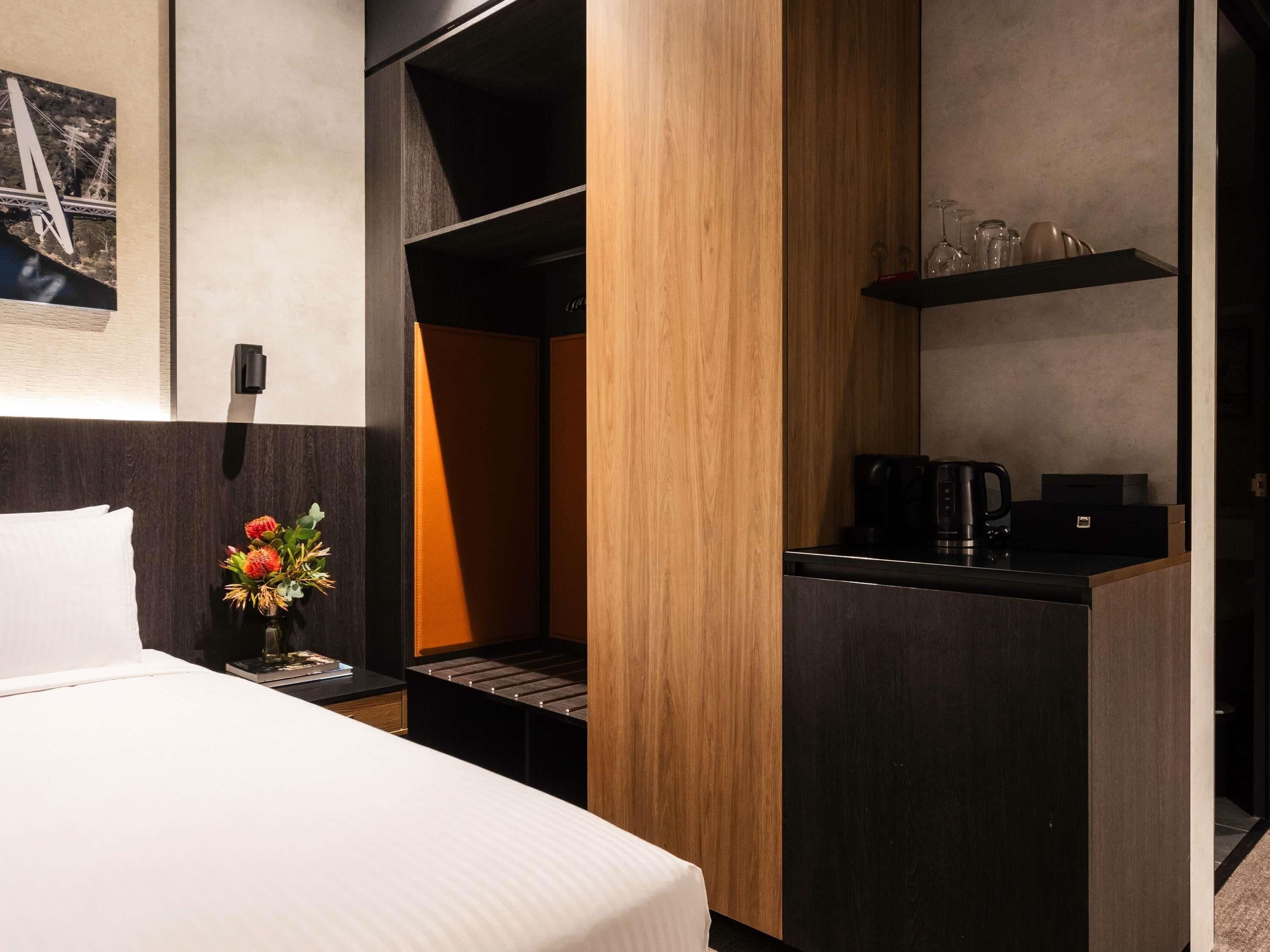 Una habitación de hotel moderna con cama, escritorio con cafetera, estante con vasos y una fotografía de un puente.